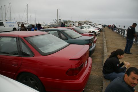 1990 Ford Taurus - Several cars are parked in a line on a wooden pier, with boats visible in the harbor.