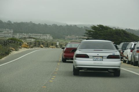 1990 Ford Taurus - A silver sedan drives along a coastal highway with parked cars on the right.