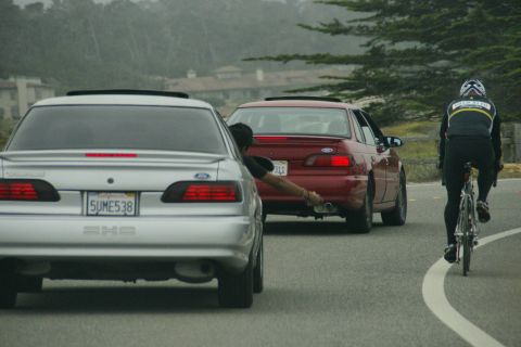 1990 Ford Taurus - A silver 1990 Ford Taurus sedan is seen from the rear, alongside a red sedan and a cyclist on the road.