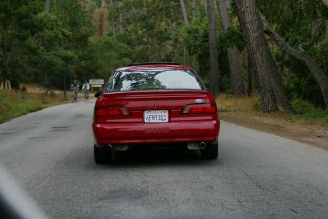 1990 Ford Taurus - Rear view of a red 1990 Ford Taurus sedan driving on a paved road with trees in the background.