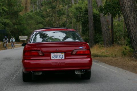 1990 Ford Taurus - Rear view of a red 1990 Ford Taurus driving on a road with trees and two cyclists in the background.