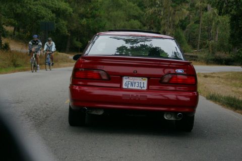 1990 Ford Taurus - A red sedan drives down a road with two cyclists in the distance.
