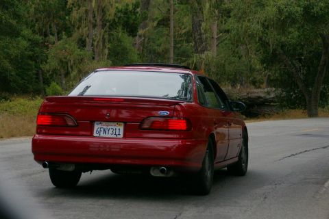 1990 Ford Taurus - A red 1990 Ford Taurus sedan is seen from the rear driving on a paved road with trees in the background.