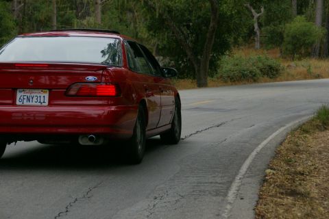 1990 Ford Taurus - A red 1990 Ford Taurus is seen from the rear driving on a winding road.