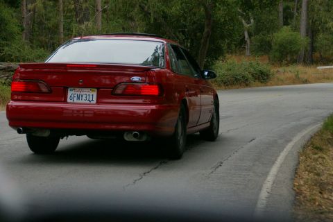 1990 Ford Taurus - Red 1990 Ford Taurus driving on a winding road with trees in the background.