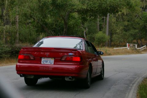 1990 Ford Taurus - A red 1990 Ford Taurus drives along a winding road bordered by trees and dry grass.