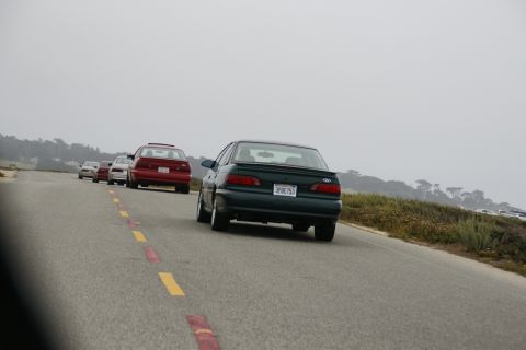1990 Ford Taurus - A line of cars, including a dark green 1990 Ford Taurus, drive along a coastal road under an overcast sky.