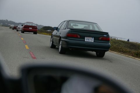 1990 Ford Taurus - A dark green sedan, possibly a 1995 Ford Taurus, drives along a coastal highway with other vehicles in the background.