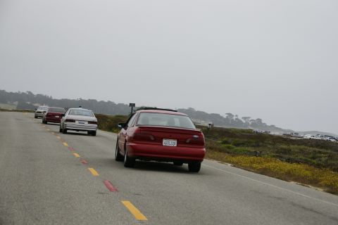 1990 Ford Taurus - Several cars drive along a scenic coastal highway under an overcast sky.