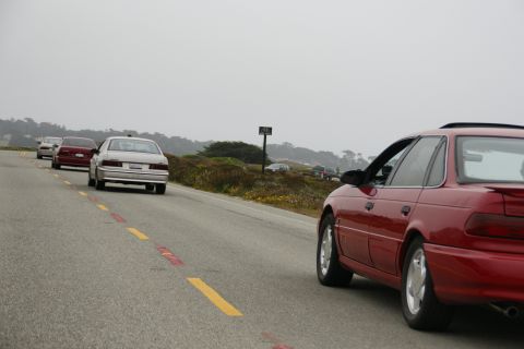 1990 Ford Taurus - A red 1990 Ford Taurus sedan drives along a coastal highway with other vehicles in the distance.