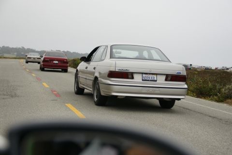 1990 Ford Taurus - Three cars driving on a coastal road, with the lead car being a 1990 Ford Taurus.