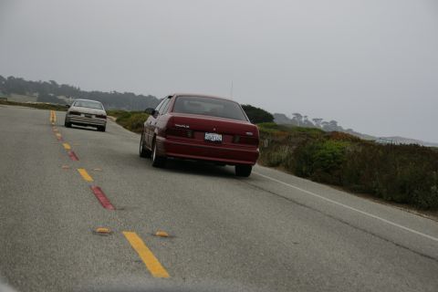 1990 Ford Taurus - Two sedans travel along a scenic coastal highway under an overcast sky.