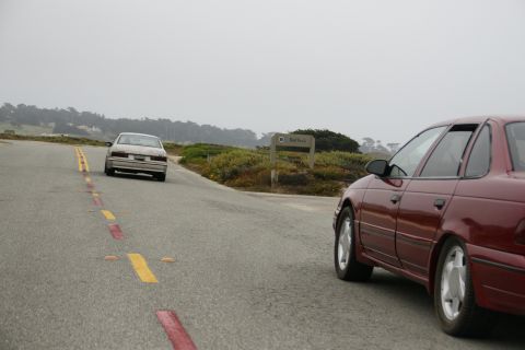 1990 Ford Taurus - Two sedans drive on a coastal road under a foggy sky, with a sign for 'Red Rock' visible.