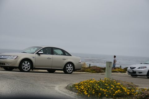 1990 Ford Taurus - A tan sedan is parked near the ocean, with a second car visible to the right. The scene suggests a coastal drive.