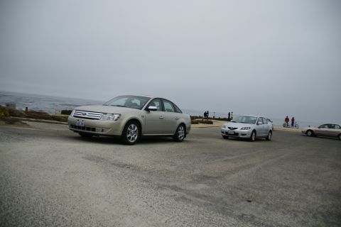 1990 Ford Taurus - A silver sedan is parked near the ocean on a cloudy day, with other vehicles and pedestrians visible.