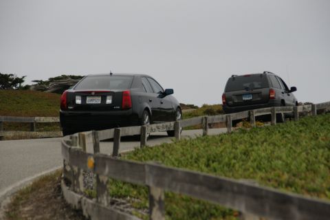 1990 Ford Taurus - Two vehicles drive along a coastal road, separated by a wooden fence.