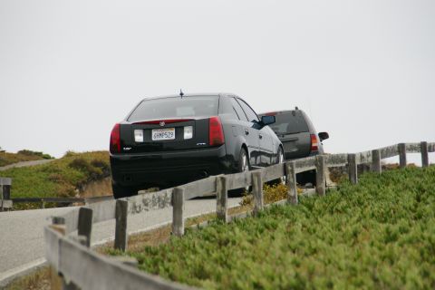1990 Ford Taurus - A black Cadillac sedan and a dark SUV drive along a coastal road, separated by a wooden fence.