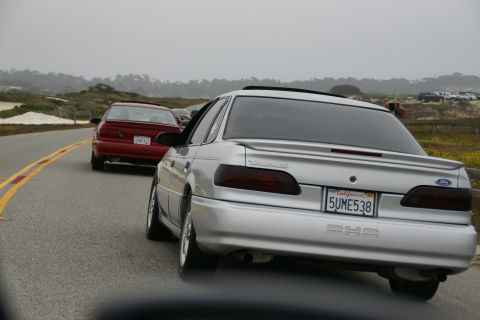 1990 Ford Taurus - A silver sedan with a spoiler and tinted taillights drives on a coastal road, with a red car ahead and a parking lot in the distance.