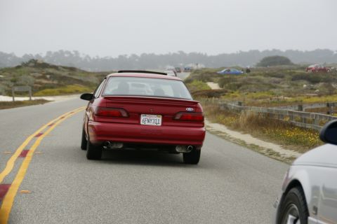 1990 Ford Taurus - A red sedan drives along a coastal highway, with another vehicle partially visible in the foreground.