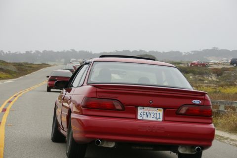 1990 Ford Taurus - A red 1990 Ford Taurus sedan is seen from the rear, driving in a line of cars on a coastal road.
