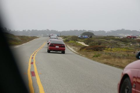 1990 Ford Taurus - A line of cars, including a red 1990 Ford Taurus, drive along a coastal highway.
