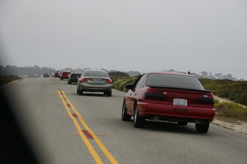 1990 Ford Taurus - A line of cars drives down a coastal road, led by a red 1990 Ford Taurus with a spoiler.