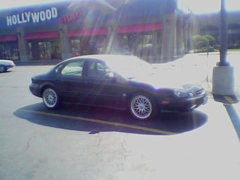 1999 Ford Taurus - A dark sedan with aftermarket wheels is parked in a lot in front of a Hollywood Video store.