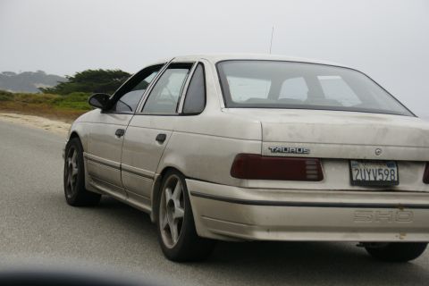 1990 Ford Taurus - Rear view of a tan 1990 Ford Taurus SHO sedan driving on a coastal road.