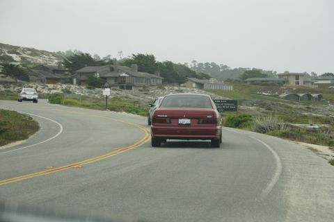 1990 Ford Taurus - A red sedan drives along a scenic coastal highway, with houses dotting the hillsides.