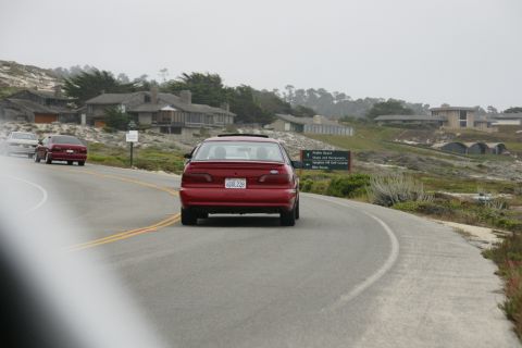 1990 Ford Taurus - A red sedan drives along a scenic coastal highway with houses and trees in the background.