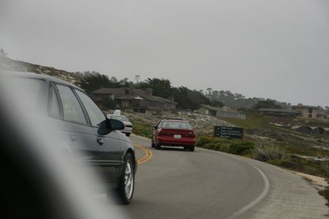 1990 Ford Taurus - A dark sedan drives along a coastal road, with a red sedan ahead and houses on a hill in the background.