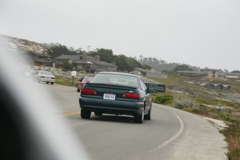 1990 Ford Taurus - A dark green 1990 Ford Taurus drives along a scenic coastal road, with other vehicles visible.