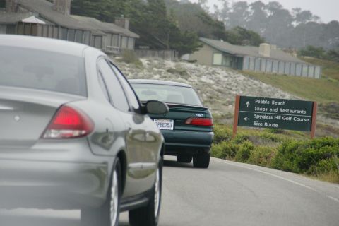 1990 Ford Taurus - Two cars drive past a sign for Pebble Beach and Spyglass Hill Golf Course on a coastal road.