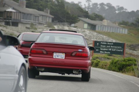1990 Ford Taurus - A red sedan with a sunroof drives along a coastal road, with a sign pointing to Pebble Beach.