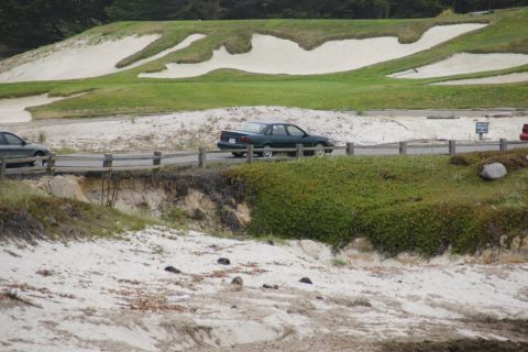 1990 Ford Taurus - A dark green 1990 Ford Taurus sedan drives along a coastal road with sand dunes and a golf course in the background.