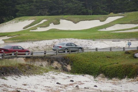 1990 Ford Taurus - Two cars drive past a golf course with sand traps and trees. The red car is on the left, silver sedan on the right.