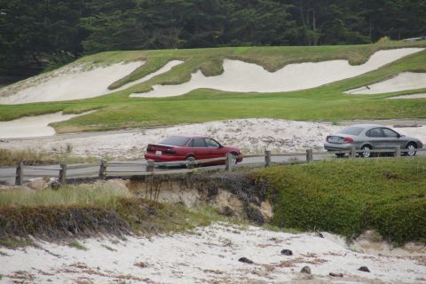 1990 Ford Taurus - Two sedans travel along a coastal road with a golf course in the background.