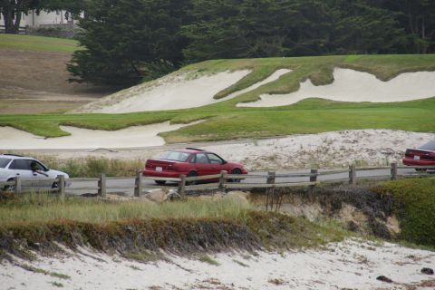 1990 Ford Taurus - A red sedan drives past a golf course's sand traps and green, with another car on the road.