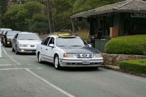 1990 Ford Taurus - A white 1990 Ford Taurus with a surfboard on its roof rack is seen in line at a gatehouse.