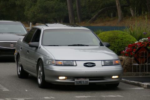1990 Ford Taurus - A silver sedan is seen from the front, with a dark car partially visible to its left. The license plate is clearly visible.