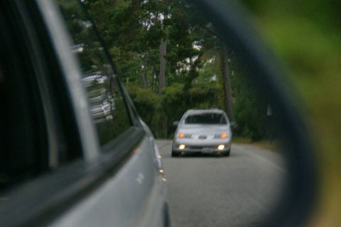 1990 Ford Taurus - A silver 1990 Ford Taurus is seen from the side mirror of another vehicle on a tree-lined road.