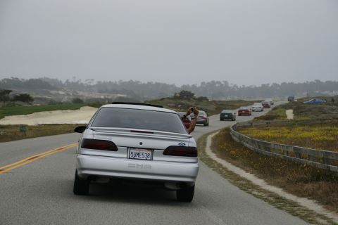 1990 Ford Taurus - A silver 1990 Ford Taurus drives along a coastal road, with other vehicles in the distance.