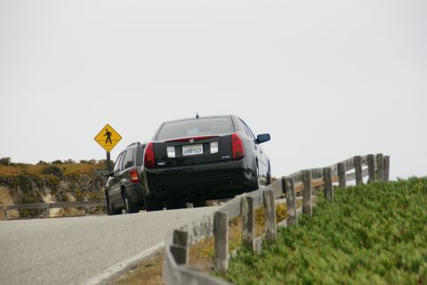 1990 Ford Taurus - Two vehicles drive along a coastal road, with a pedestrian crossing sign visible.