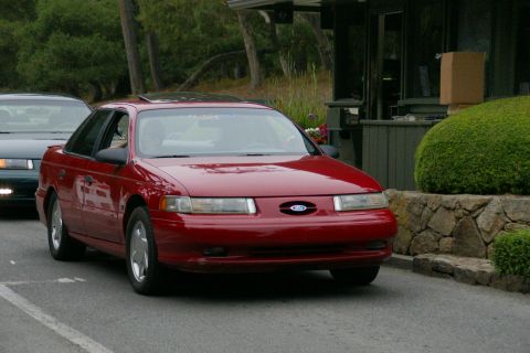 1990 Ford Taurus - A red 1990 Ford Taurus sedan is seen driving on a road, with another vehicle partially visible in the background.