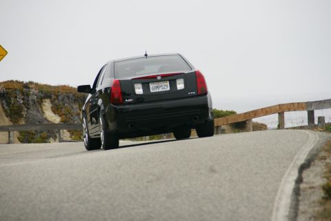1990 Ford Taurus - A black Cadillac CTS navigates a winding road along a coastline under an overcast sky.