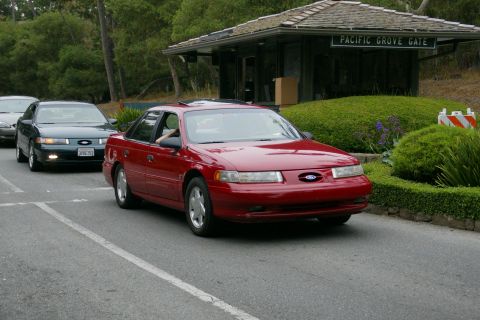 1990 Ford Taurus - A red 1990 Ford Taurus is seen driving past the Pacific Grove Gate, with another car following behind.
