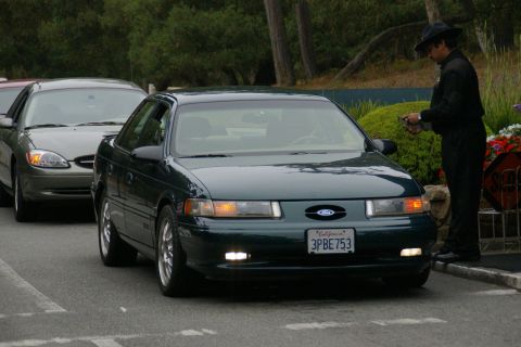 1990 Ford Taurus - A dark green 1990 Ford Taurus is parked on the street, with a man in a hat standing beside it. Another car is visible behind it.