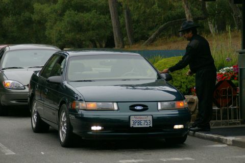 1990 Ford Taurus - A man in a uniform interacts with a dark green sedan, with another vehicle visible behind it.