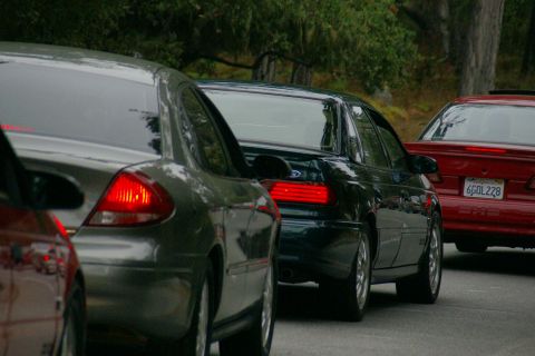 1990 Ford Taurus - A line of cars are driving on a road with trees in the background. The license plate on the red car is visible.