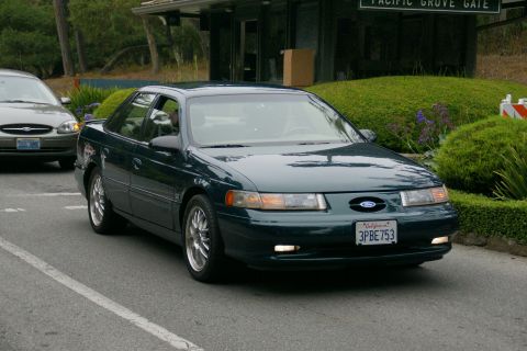 1990 Ford Taurus - A dark green 1996 Ford Taurus drives past a gate, with another car visible behind it.